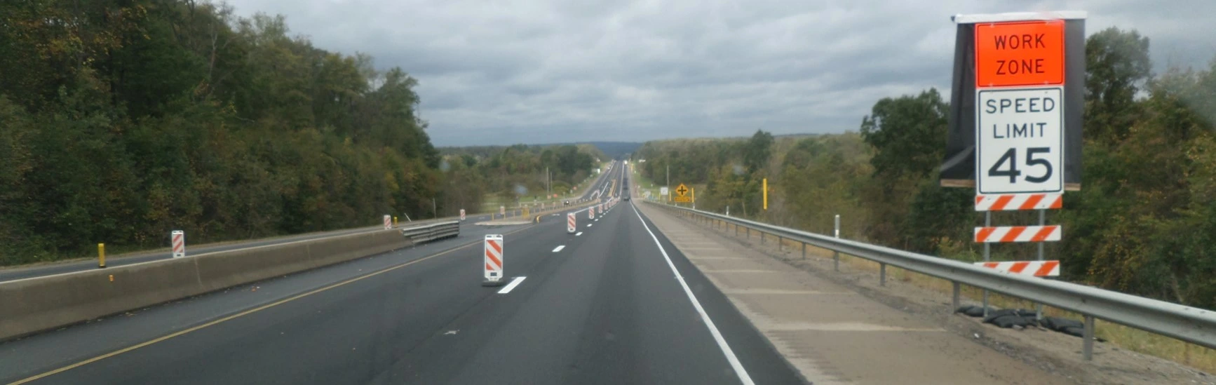 An image showing a blacktop highway with an orange work-zone sign and a white speed limit sign with 45 miles per hour in black letters and numbers, and trees on either side of the road.